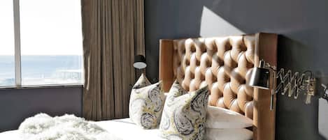 A bedroom with a tufted headboard, a bedside lamp, patterned pillows, a fluffy throw, and a view of the city through a window.