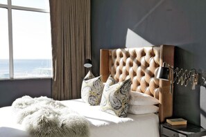 A bedroom with a tufted headboard, a bedside lamp, patterned pillows, a fluffy throw, and a view of the city through a window. - Stylish Clifton Apartment - Number 5102 (Cape Town)
