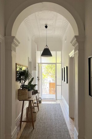 Um corredor com teto em arco, luminária pendente preta, um banco de madeira, plantas em vasos e quadros nas paredes.