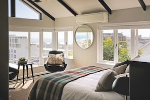 A bedroom with a bed, a chair, a small table, and a large window with a view of buildings and greenery.