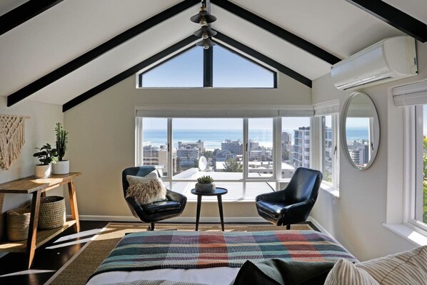 A high-ceiling room with a large window, two black leather chairs, a small table, and a bed with a colorful blanket.