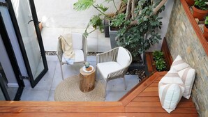 A wooden deck with white cushioned chairs and a small round table, surrounded by potted plants and a stone wall.