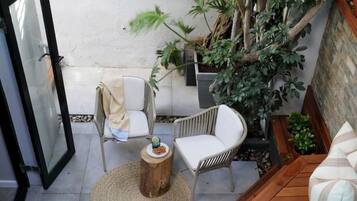 A poolside area with a wooden deck, a white outdoor sofa, a wicker chair, and a small round table with a plate of snacks.