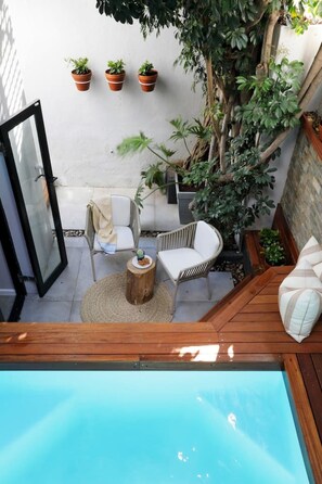 A poolside area with a wooden deck, a white outdoor sofa, a wicker chair, and a small round table with a plate of snacks.