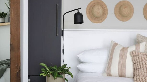 Een slaapkamer met een bed, een nachtkastje met een plant, een wandlamp en een deur.