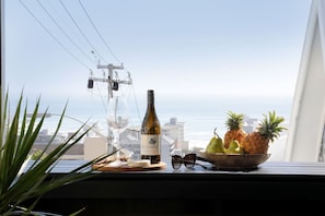 A bottle of wine, a bowl of fruit, and a pair of glasses on a windowsill with a view of the ocean and buildings.
