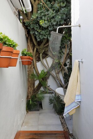 A narrow outdoor space with a white wall, a wooden floor, a hanging shower, and potted plants.