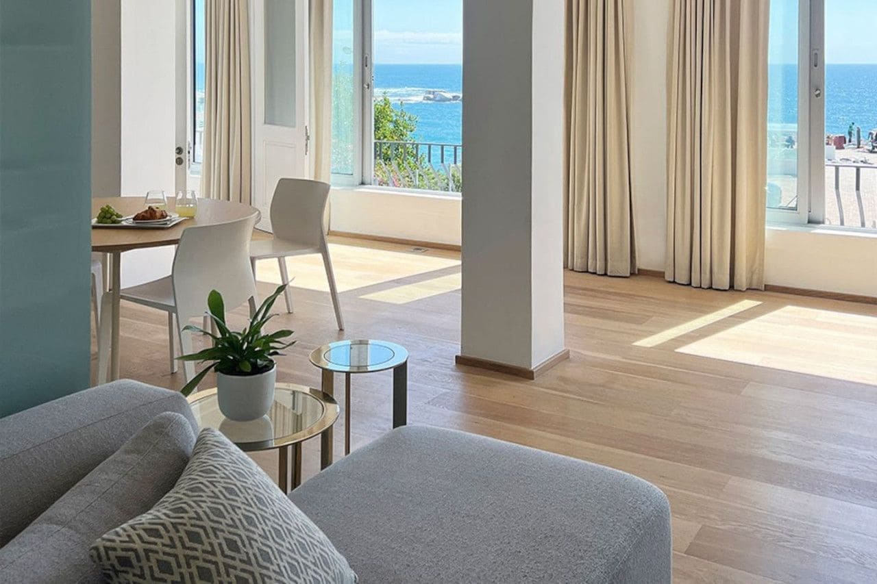 A modern living room with a view of the sea, a dining area with a table and chairs, and a balcony with curtains.