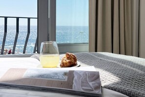 A hotel room with a view of the ocean, a glass of water, a pastry, and a magazine on the bed.