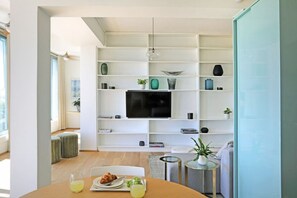 A modern living room with a flat-screen TV, white shelving, and a dining area with a table set for two.