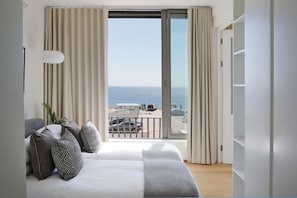 A bedroom with a bed, pillows, a bedside table, and a view of the sea through a balcony.