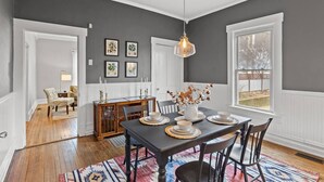 Un coin repas avec une table en bois foncé, dressée pour quatre personnes et entourée de chaises. Sur la table, on trouve un vase contenant des fleurs séchées, et un luminaire suspendu au-dessus. Les murs sont peints en gris, et des reproductions botaniques encadrées ornent les murs.