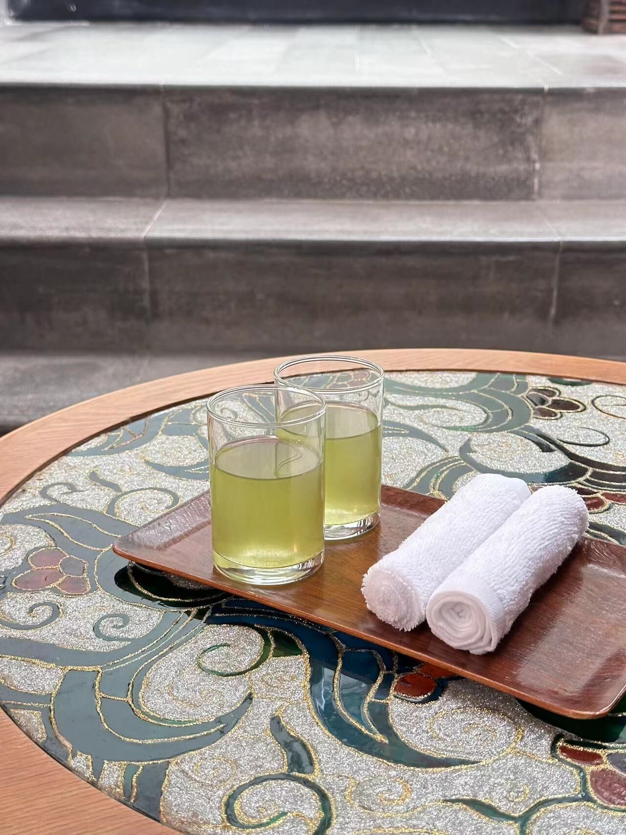 Una mesa redonda con una bandeja decorativa que sostiene dos vasos de una bebida amarillenta y una toalla blanca enrollada.