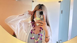 Une personne prend un autoportrait dans une chambre d’hôtel, en utilisant un miroir. On aperçoit un plafond blanc, une bouche d’aération et une porte bleu clair.