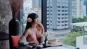Une femme est assise à une table, un verre à la main, et contemple un paysage urbain dominé par de hauts immeubles.