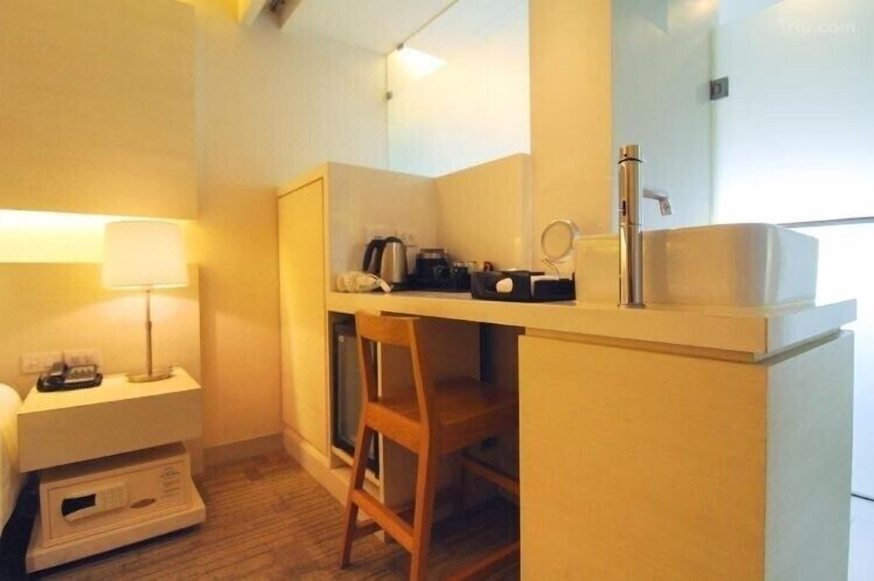Une chambre d’hôtel de taille modeste, comprenant un petit bureau, une chaise, une table de chevet avec une lampe, un miroir et un lavabo.