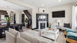 A modern living room with a fireplace, a dining area, and a staircase leading to another level. - Stylish 4BR Home • Close to Hospitals + Downtown (Cleveland)