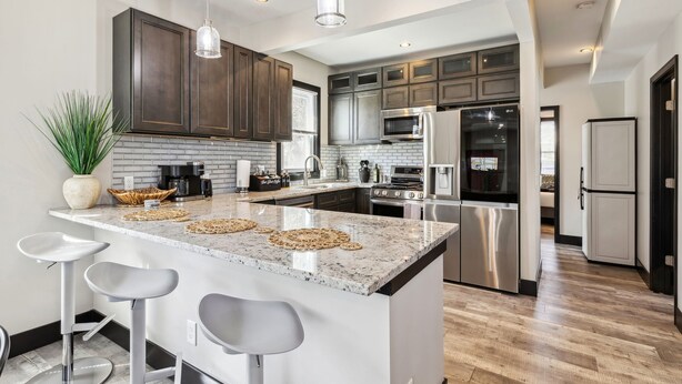 A modern kitchen with dark wood cabinets, a granite island with bar stools, stainless steel appliances, and pendant lighting. - Stylish 4BR Home • Close to Hospitals + Downtown (Cleveland)