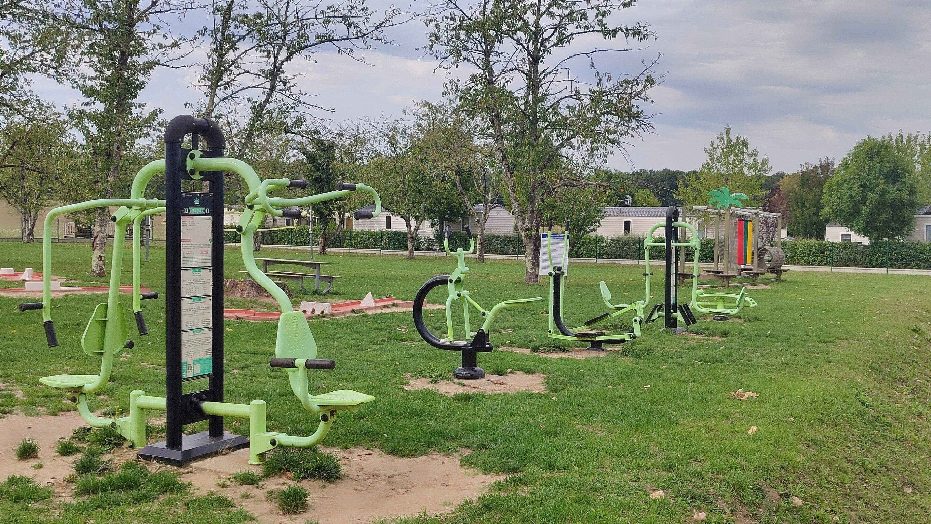 Draußen stehende Fitnessgeräte in einem Park mit Rasen, Bäumen und einer Bank.
