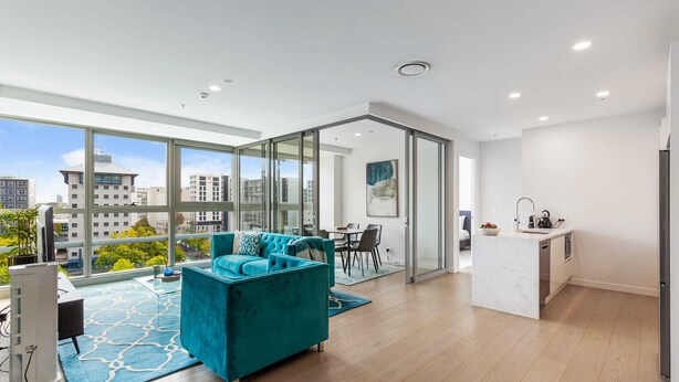 Un appartement moderne doté d’une grande porte-fenêtre coulissante donnant sur un balcon, d’un canapé de couleur turquoise, d’un coin cuisine et offrant une vue sur la ville.