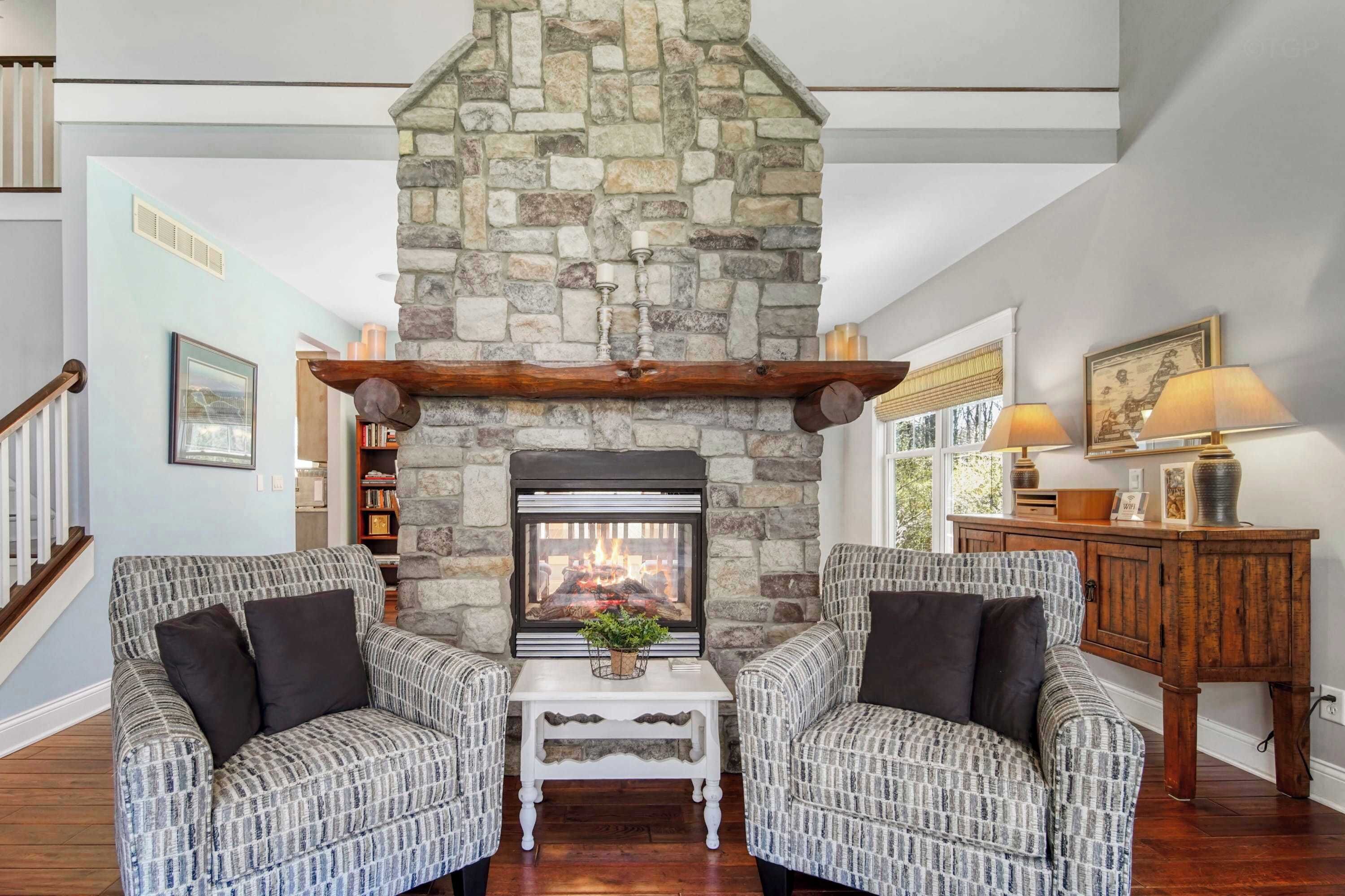 A cozy living room with a stone fireplace, two patterned armchairs, a small white table, and a wooden cabinet with a lamp.