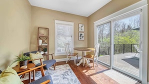 Un salon confortable avec un canapé, des fauteuils, une table basse et une bibliothèque. Il y a une grande fenêtre avec des stores, une porte donnant sur un balcon et une vue sur les arbres.