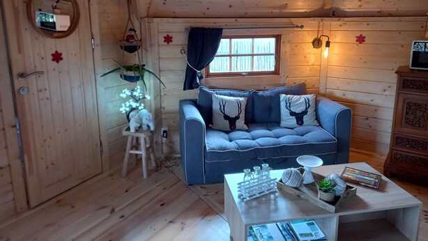 Et hyggeligt sommerhusindretning med en blå sofa, trævægge, en lille skammel, et bord med pyngeting og et vindue med gardiner.
