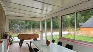 Et moderne spiseområde med et stort bord og stole, store vinduer der giver udsigt over en have og en trækonstruktion.