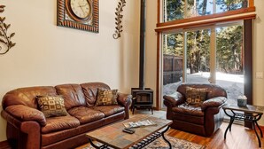 A spacious living room with a brown leather sofa, a coffee table, and a large window offering a view of trees. - Golfers View - on Golf Course - Close to park and lake (McCall)