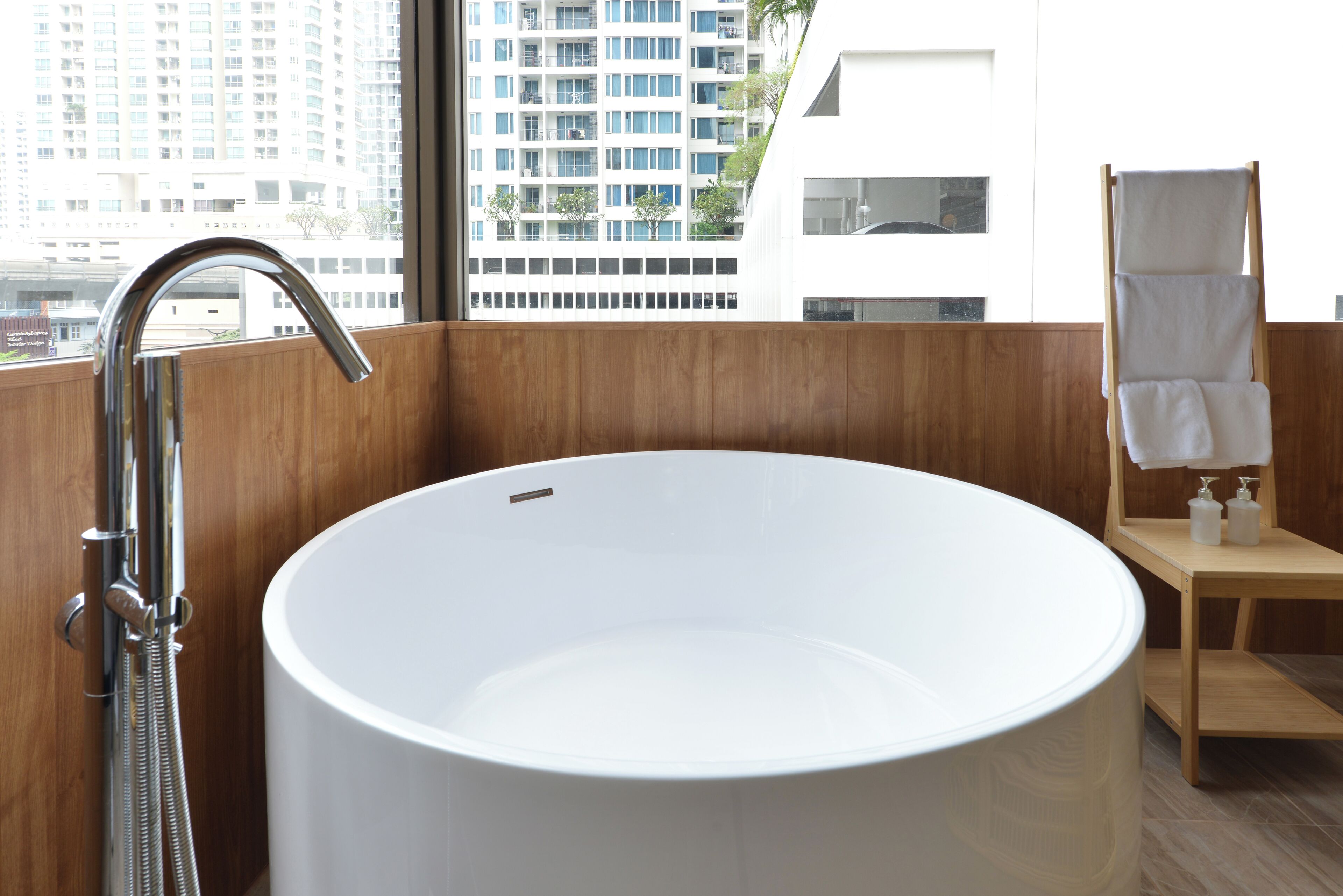 A modern bathroom with a large white bathtub, wooden paneling, and a view of tall buildings through a large window.