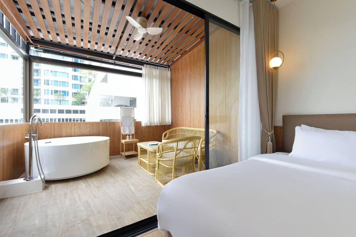 A modern hotel room with a large bed, a bathtub, wicker furniture, and a city view.