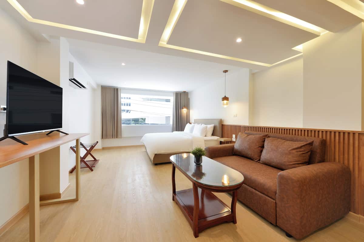 A modern hotel room with a flat-screen TV, a wooden desk, a brown sofa, a glass coffee table, and a bed with white linens.