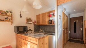 Eine kleine Küche mit Holzausstattung, einer Mikrowelle, einem Spülbecken und einem Kühlschrank. Eine Holztür führt in einen weiteren Raum.