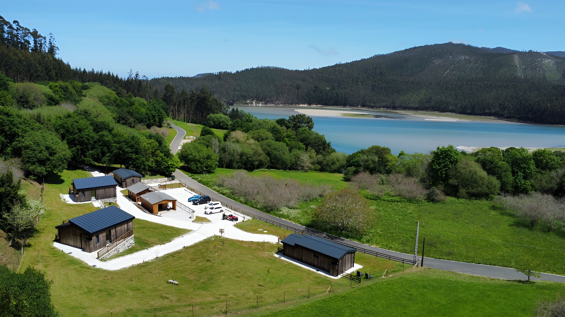 Una propiedad rural con varias cabañas, un estacionamiento y una vista panorámica de un lago y colinas boscosas.