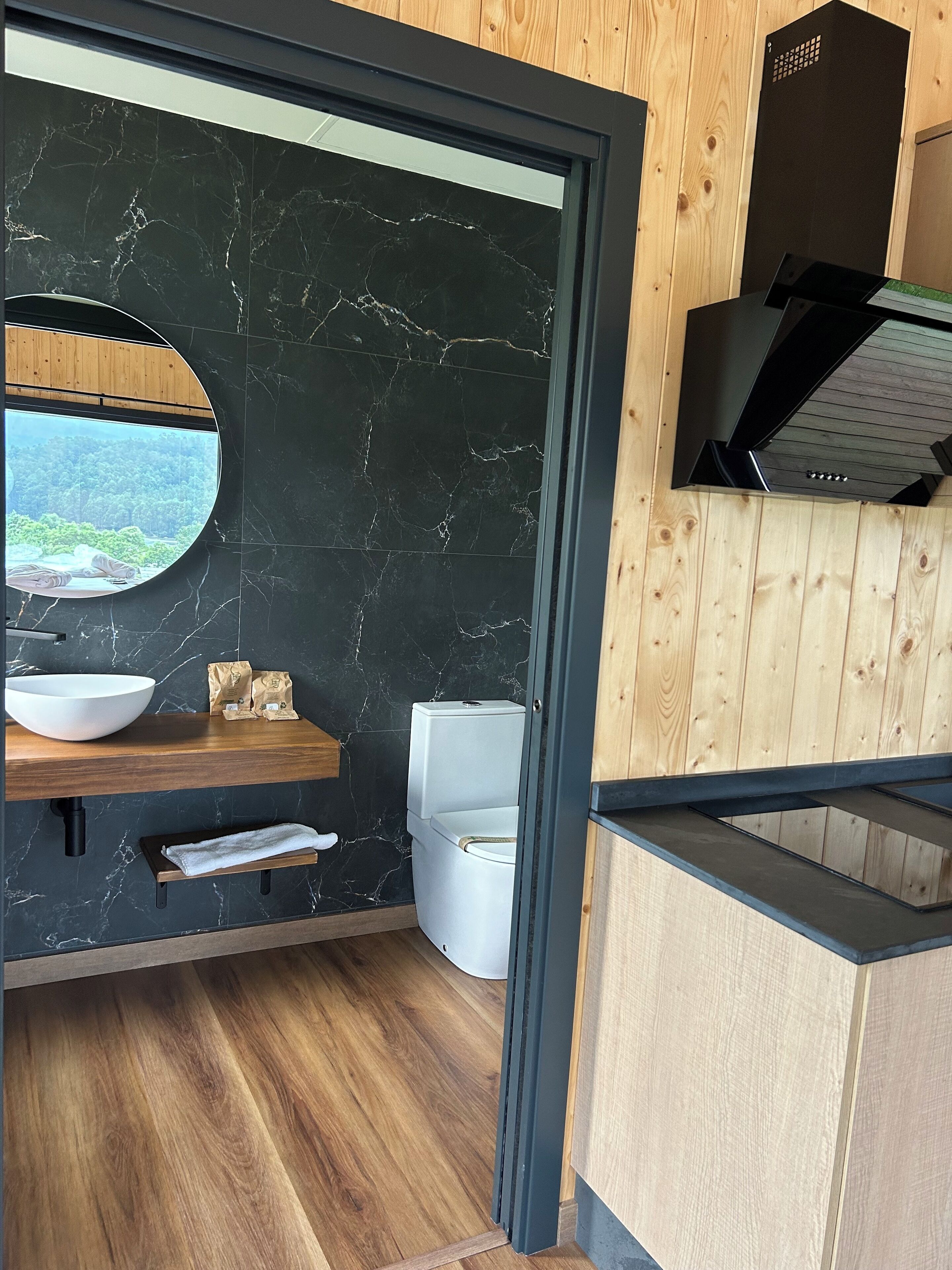 A modern bathroom with a wooden vanity, a large mirror, a white toilet, and a view of a forest through a circular window.