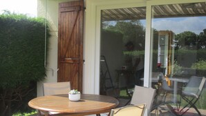 A patio with a round wooden table and two chairs, a sliding glass door, and a wooden door. - Gîte le Lilas (vierville sur mer  France)