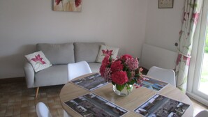 A living room with a grey sofa, a coffee table with photographs, a vase with flowers, and a window with curtains. - Gîte le Lilas (vierville sur mer  France)