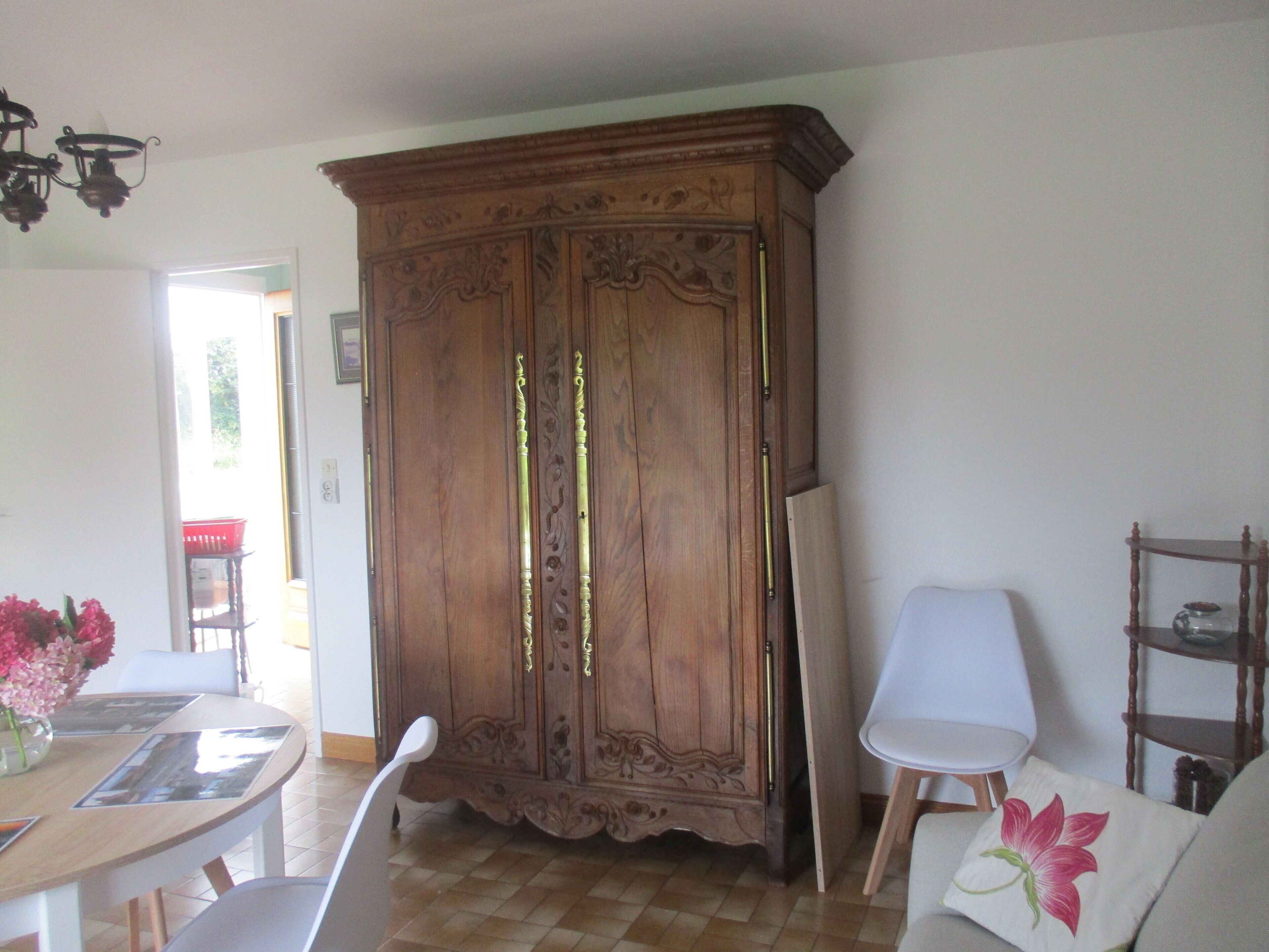 A room with a wooden wardrobe, a dining table with chairs, a white armchair, and a small wooden shelf.
