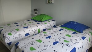 A room with two single beds, each with a patterned bedspread, a green pillow on one bed, and a blue pillow on the other. There is a small bedside table with a lamp and a shelf with books and decorative items. - Gîte le Lilas (vierville sur mer  France)