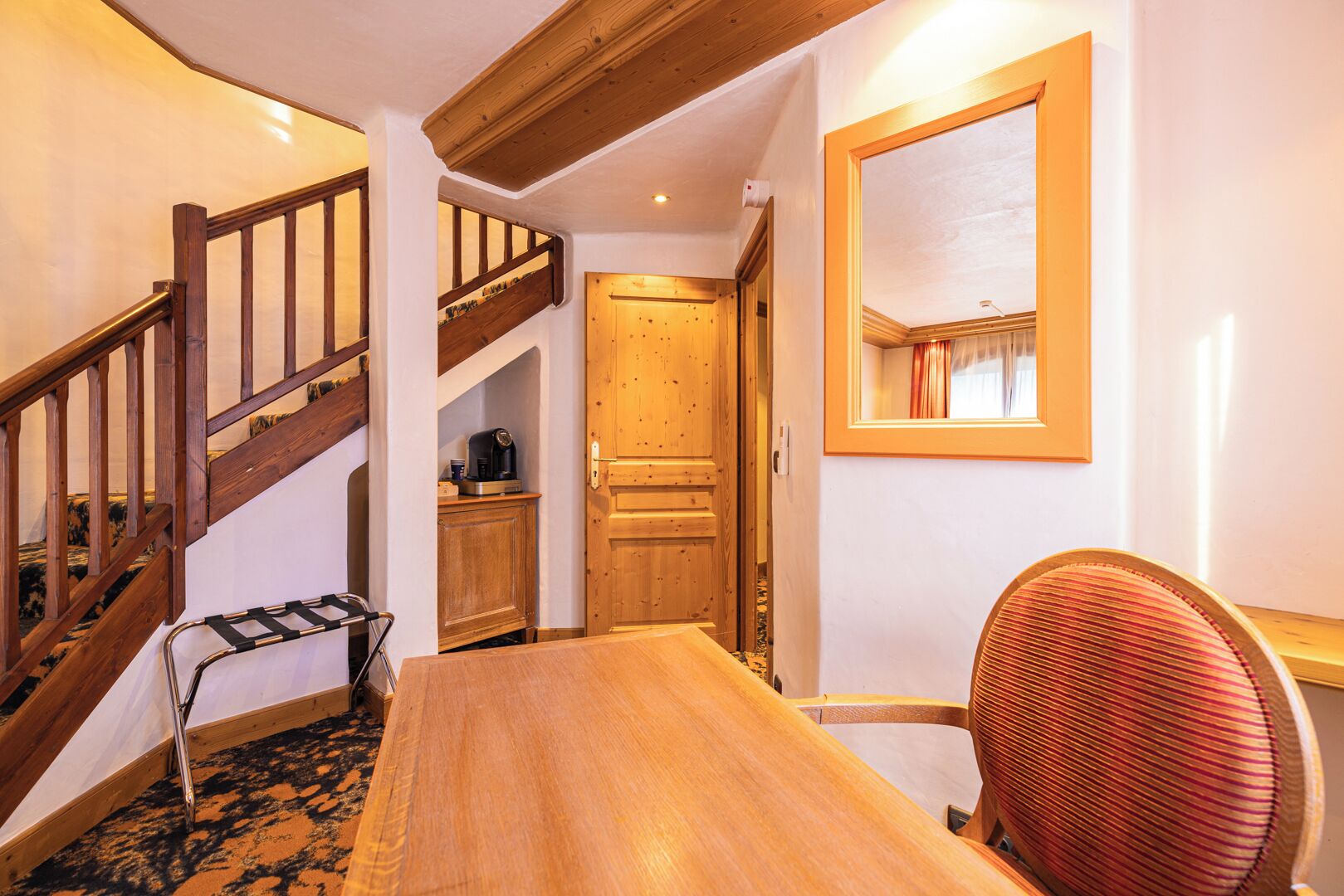 Una mesa de comedor de madera con una silla, una escalera con barandilla de madera y una puerta con un espejo encima.