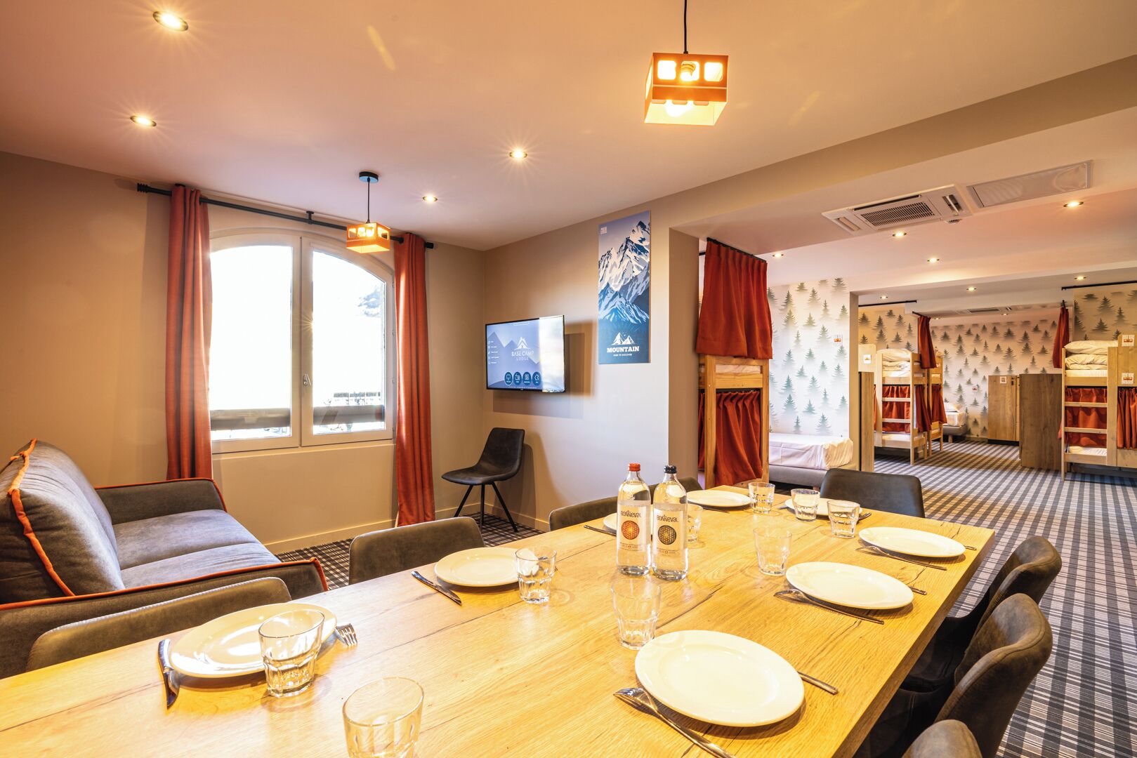 A hotel room with a dining table set for four, a sofa, a TV, and a mountain-themed wall.