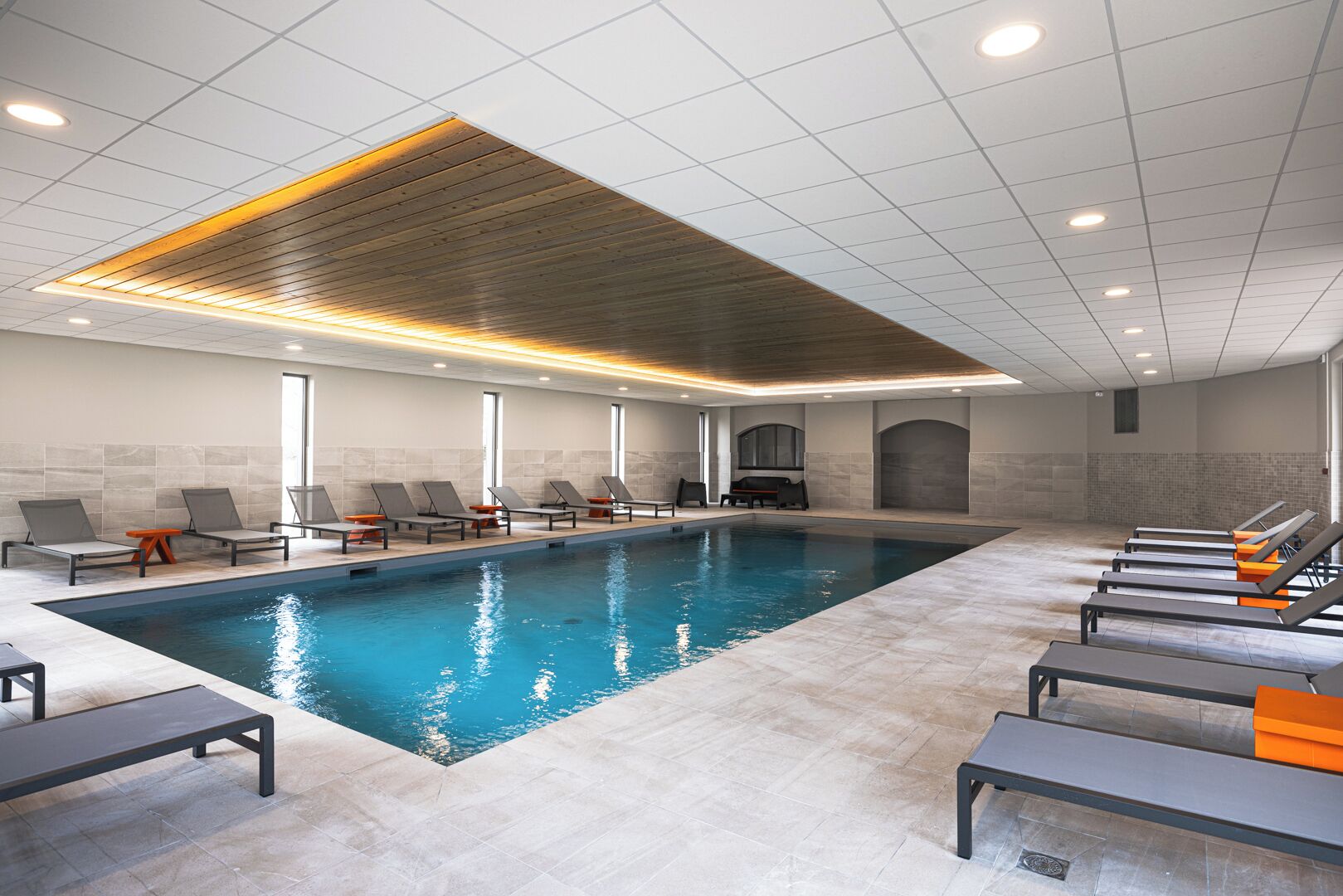 A modern indoor swimming pool with lounge chairs and a wooden ceiling.