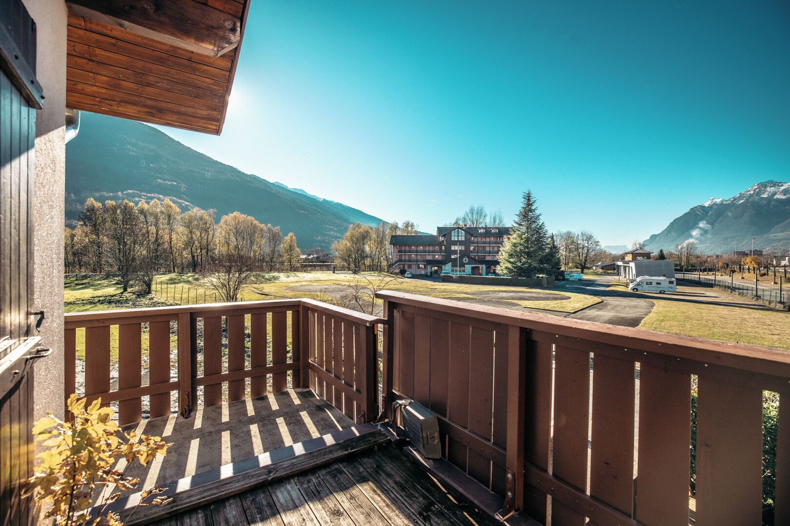 A wooden deck with a view of a mountainous landscape, a building with multiple windows, and a parked van.