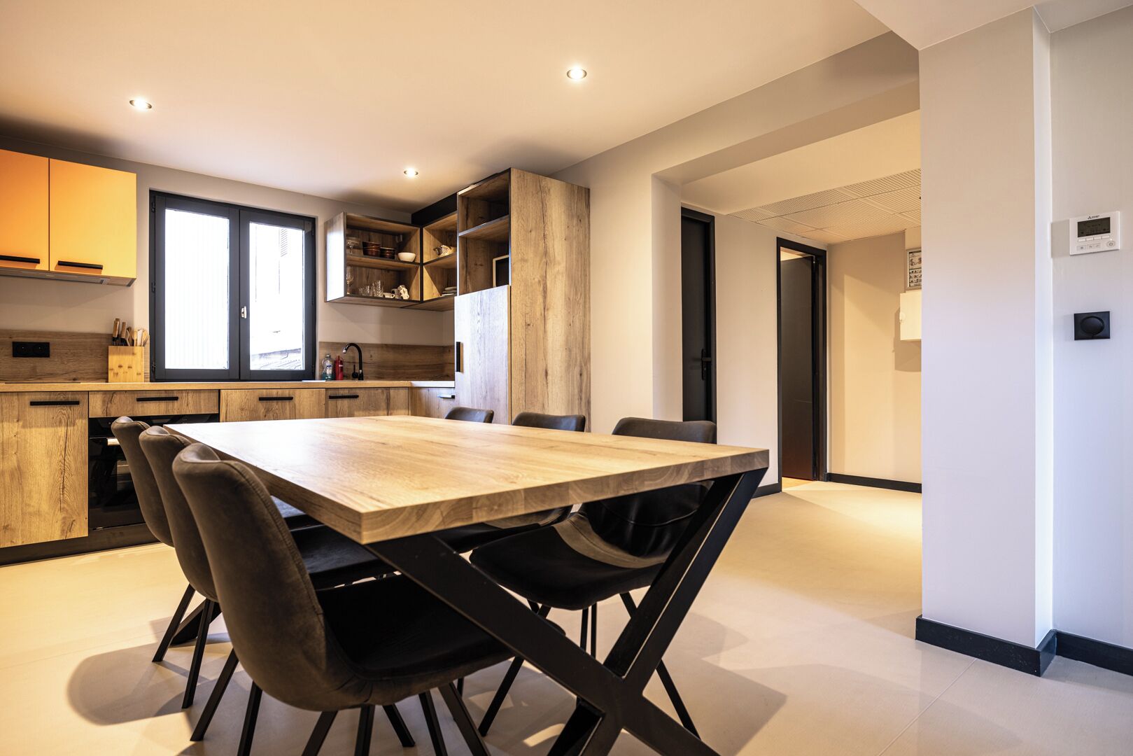 A modern kitchen with a wooden table and chairs, built-in shelves, and a large window.