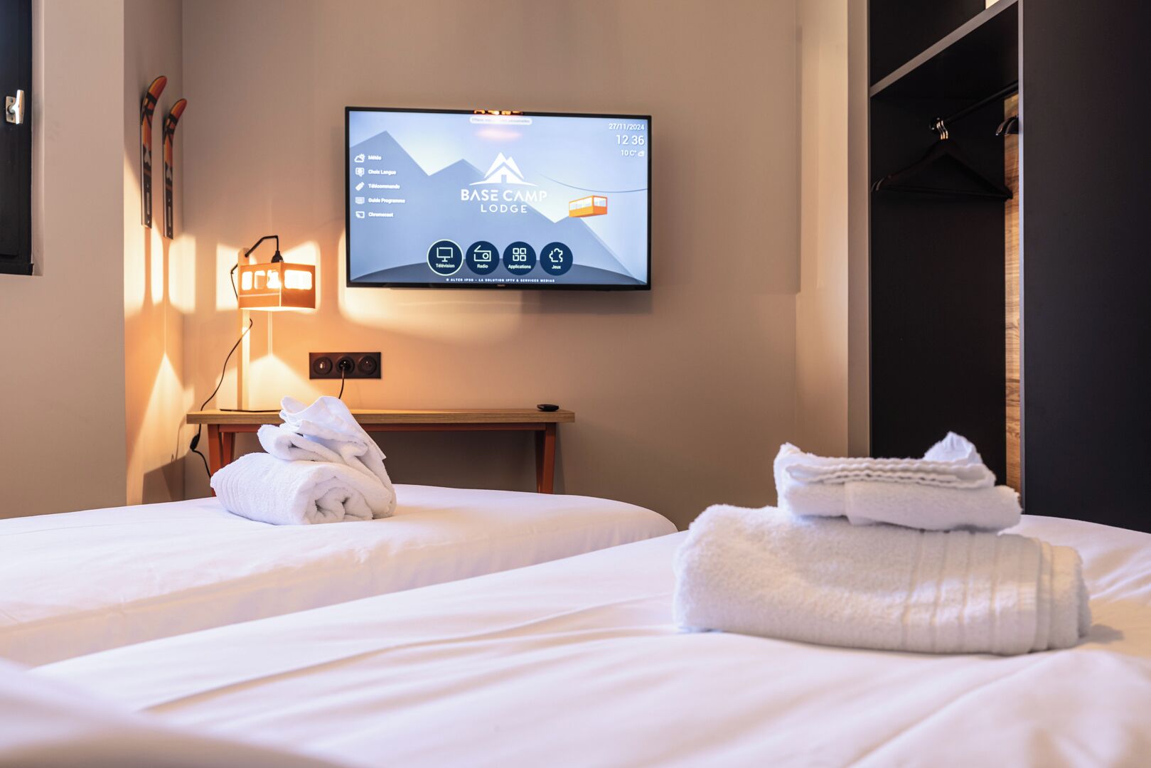 A hotel room with two beds, a TV displaying the BASE CAMP LODGE interface, and a ski rack against the wall.