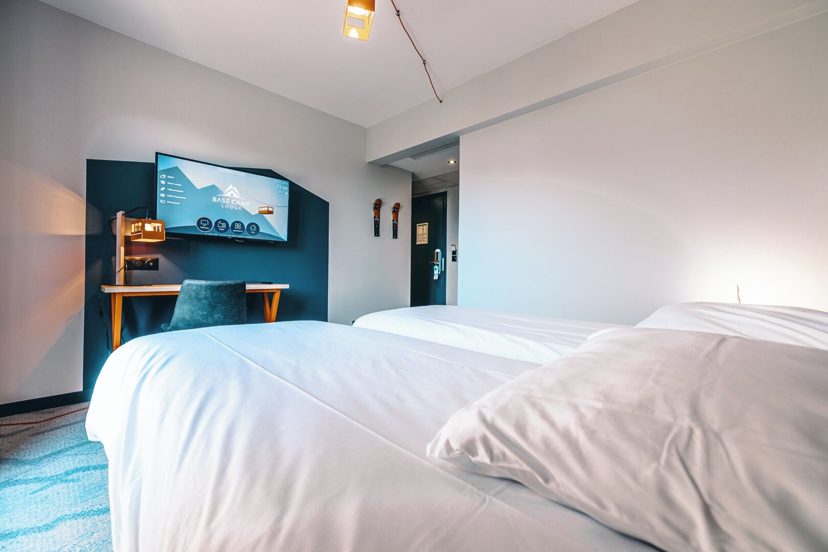 Habitación de hotel con dos camas, un escritorio con silla, un televisor montado en la pared y una puerta que da a otra habitación.