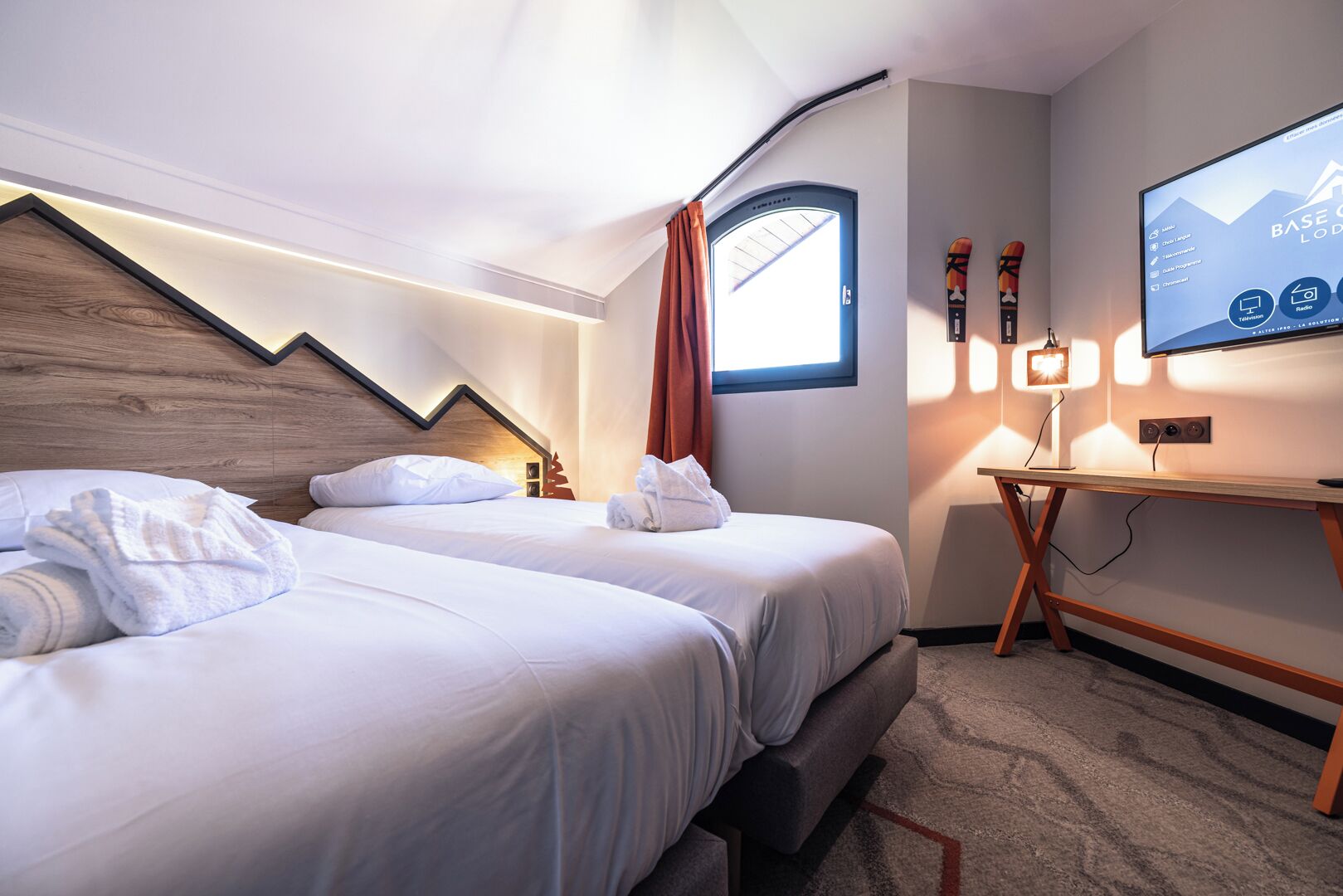 A modern hotel room with two beds, a desk, and a TV displaying a mountain landscape.