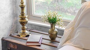 A bed with white linens, a brass lamp, a vase with flowers, and a vintage suitcase with books. - Vintage Veranda - Ping Pong Table & More! (Fredericksburg)