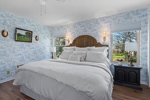 Une chambre avec un grand lit, une tête de lit en bois, deux lampes fixées au mur, une table de chevet et une fenêtre donnant sur un paysage verdoyant.