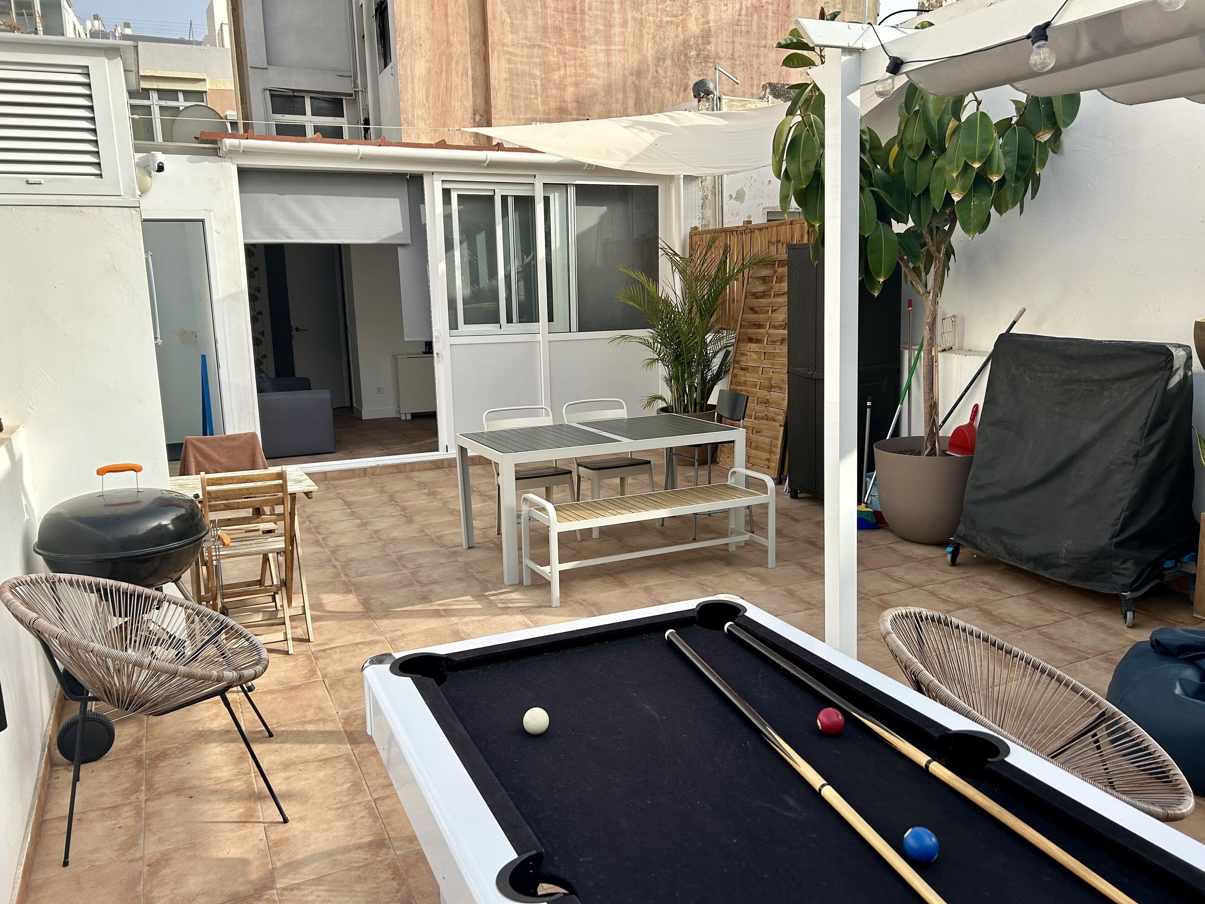 Mesa de billar con bolas, una parrilla, asientos al aire libre y una zona cubierta con plantas.